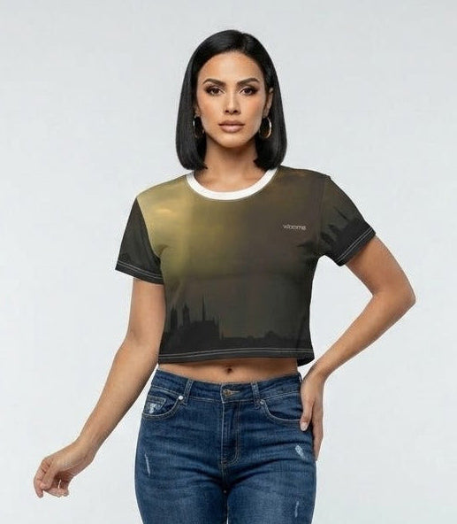 A woman modeling a short-sleeve crop top with a dark olive and black city skyline print, paired with dark blue skinny jeans, against a light gray studio background.
