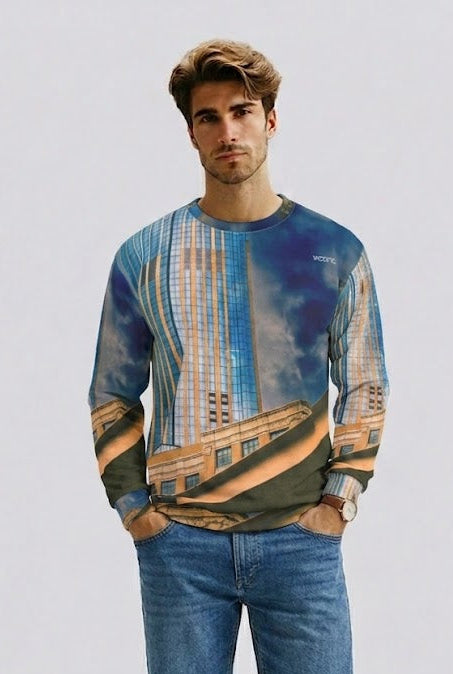 A man modeling a long-sleeve crewneck sweatshirt with a blue and tan architectural cityscape print, paired with blue denim shorts and brown sandals, against a light gray studio background.