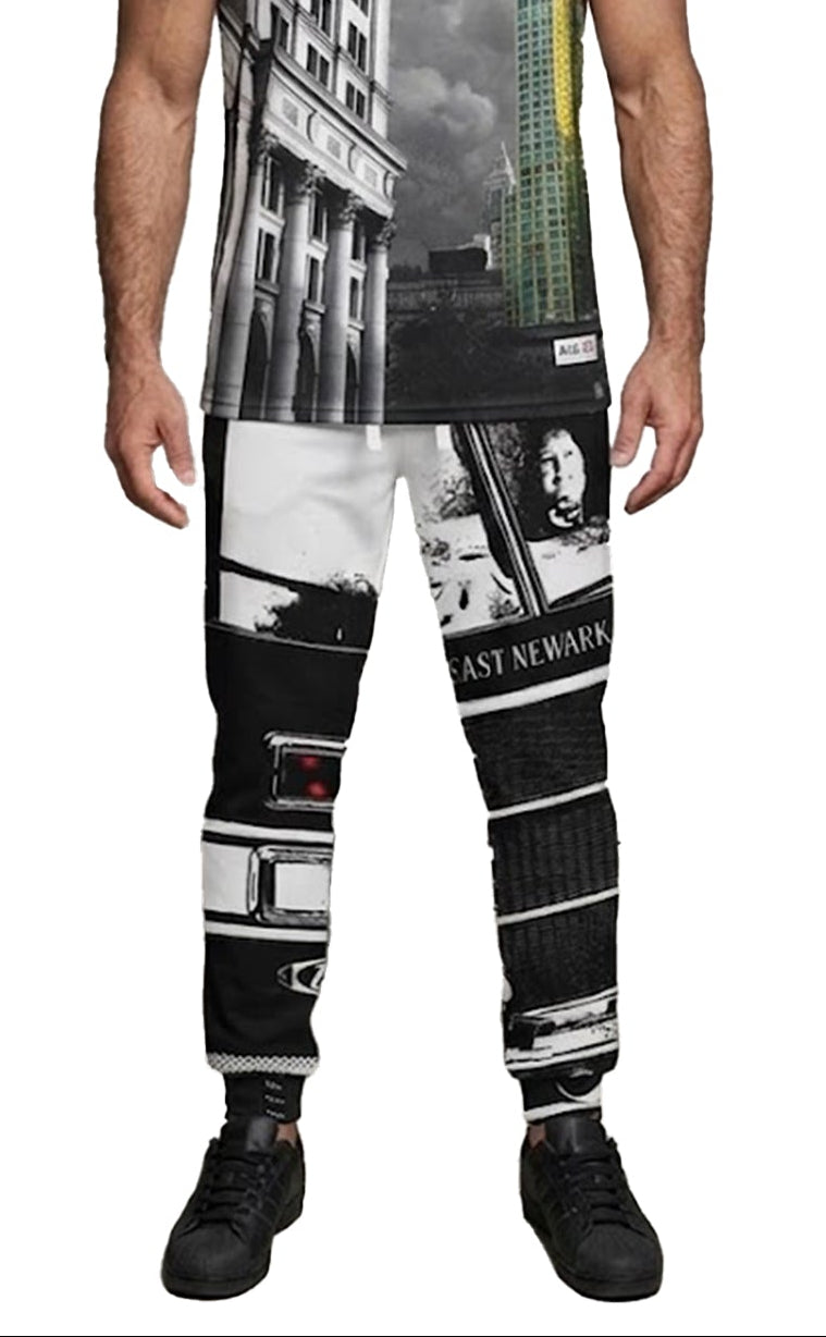 A muscular man with a graying beard stands against a white background wearing a sleeveless tank top and matching jogger pants with a black-and-white cityscape print featuring buildings, street scenes, and text.