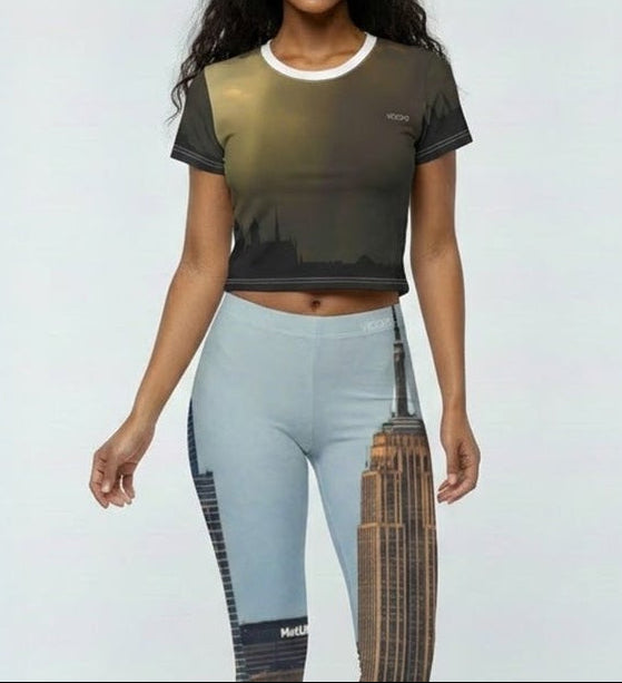 A woman stands against a light gray studio background wearing a cropped short-sleeve t-shirt with a dark olive-to-brown gradient and matching high-waisted leggings featuring a split design of light blue on one leg and a brown-and-gold abstract print on the other.