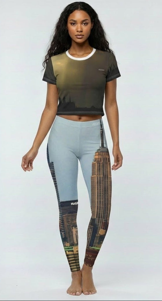 A woman stands against a light gray studio background wearing a cropped short-sleeve t-shirt with a dark olive-to-brown gradient and matching high-waisted leggings featuring a split design of light blue on one leg and a brown-and-gold abstract print on the other.